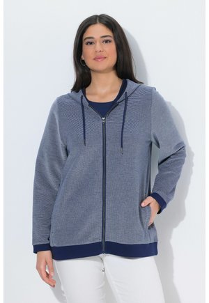 Mujer joven que lleva una sudadera con capucha y cremallera azul con cordones y pantalones blancos, de pie con una mano en el bolsillo contra un fondo liso.