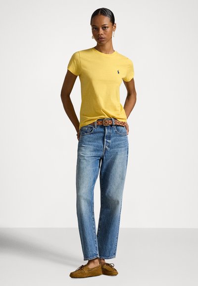 Ženska stoji oblečena v rumeno kratko majico, modre kavbojke z vtkano rjavo pasom in rjave čevlje v stilu mokasinov pred belim ozadjem.