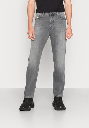 Mann trägt ausgebleichte graue Jeans mit geradem Bein, schwarze Sneakers und ein schwarzes Hemd, steht vor einfachem hellem Hintergrund.