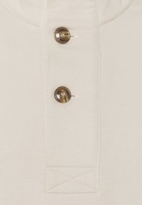 Zbliżenie na beżową tkaninę z dwoma brązowymi, marmurkowymi guzikami oraz widocznymi detalami szwów na listwie koszuli.