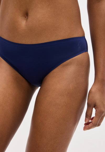 Γυναίκα με μαύρο μπλε κάτω μέρος μπικίνι, εμφανίζοντας την κοιλιακή περιοχή, δεξί χέρι χαλαρό δίπλα στο σώμα, απλό φωτεινό φόντο.