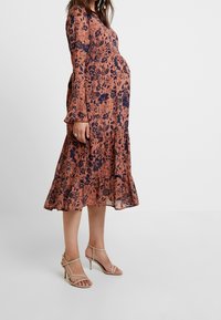 Gravid kvinna som står i en långärmad, knä-lång blommig klänning med nude-färgade sandaler på en enkel ljus bakgrund.