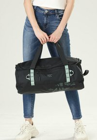 Sac de sport noir avec des accents vert menthe, en tissu robuste, avec des compartiments zippés et des poignées doubles pour le transport. Dimensions non spécifiées.
