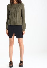 Pull en maille torsadée vert olive avec poignets côtelés, associé à une jupe en denim noir avec une fente avant et un ourlet effiloché. Chaussures marron à lacets.