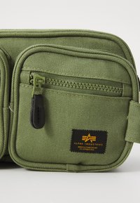 Πράσινη υφασμάτινη τσάντα με πολλαπλές τσέπες με φερμουάρ, υφή επιφάνειας και ορθογώνιο σχήμα. Διαθέτει ετικέτα "Alpha Industries" και μαύρες λεπτομέρειες φερμουάρ.