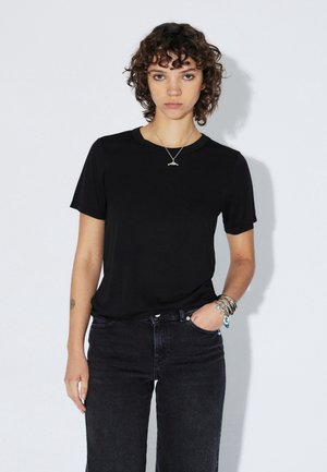 Nuori nainen, jolla on kiharat ruskeat hiukset, pukeutuneena mustaan t-paitaan, tummiin farkkuihin, hopeiseen kaulakoruun ja moniin rannekoruihin, seisoo valkoista taustaa vasten.