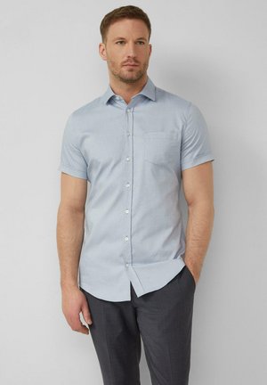 Man draagt lichtblauw overhemd met korte mouwen en borstzakje, donkergrijze broek, staat voor een effen lichtgrijze achtergrond.