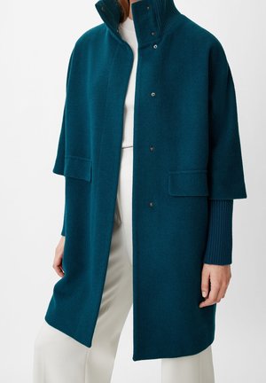 Femme portant un manteau en laine bleu sarcelle boutonné à manches courtes superposé à un pull côtelé à manches longues et un pantalon blanc.