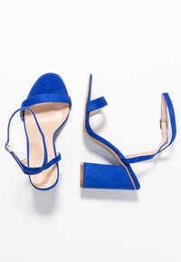 Siniset mokkanahkakengät, joissa on avonainen varvas, nilkkaremmi ja paksu lohkarese et. Niissä on sileä nude sisäpohja ja minimalistinen muotoilu.