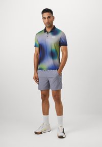 Polo shirt in gradient blue and green with a logo on the chest, paired with light gray shorts and white sneakers.