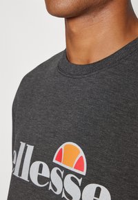 Tamno siva dukserica s okruglim izrezom, s velikim bijelim otisnutim logotipom i kružnom narančastom i crvenom grafikom iznad njega.