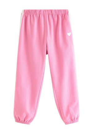 Jong meisje met krullend haar dat een roze eenhoorntrui, roze trainingsbroek met witte strepen en roze schoenen draagt en op één voet staat.