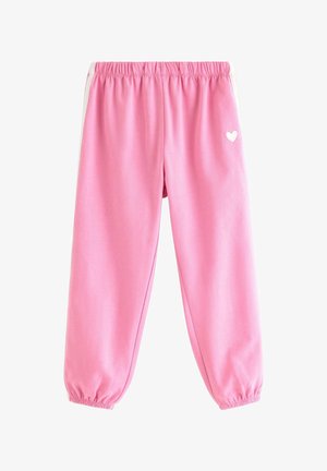 Junges Mädchen mit lockigem Haar, das einen rosa Einhorn-Sweatshirt, rosa Jogginghose mit weißen Streifen und rosa Schuhe trägt und auf einem Fuß steht.