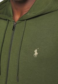 Olivově zelená mikina na zip vyrobená z měkké tkaniny, s kapucí na šňůrku a malým vyšitým logem na hrudi.