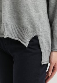 Suéter gris de punto con dobladillo acanalado, que presenta un diseño más corto por delante y más largo por detrás con aberturas laterales. La tela parece suave con una textura media. Lleva puesto sobre pantalones oscuros.