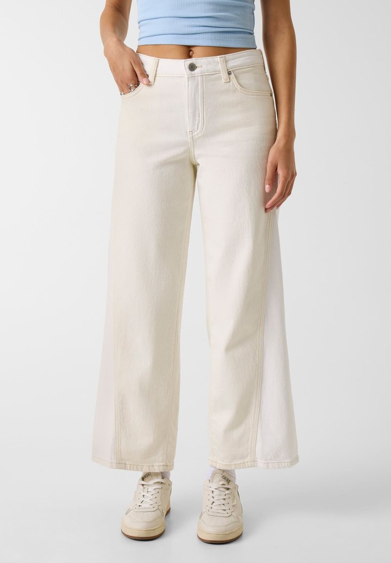 Kvinde iført cremefarvede bukser med vide ben, lyseblå cropped top og cremefarvede sneakers, der står mod en hvid baggrund.