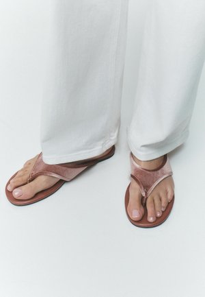 Nohy v růžových sametových žabkách ve tvaru tanga v kombinaci s bílými kalhotami na jednobarevném světlém pozadí.