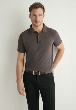 Mężczyzna stojący przy białej ścianie, ubrany w brązową koszulkę polo, czarne spodnie i brązowy pasek, patrzący prosto w kamerę.