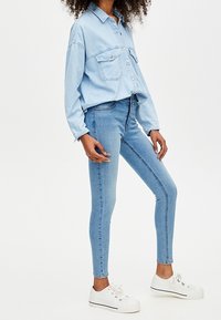 Camisa de denim azul claro con dos bolsillos en el pecho, combinada con jeans ajustados de denim azul claro. Zapatillas blancas con rayas negras. Textura suave.