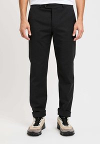 Pantaloni neri su misura realizzati in tessuto texture, con chiusura a bottone e tasche anteriori, indossati con risvolti e scarpe sportive.