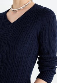 Person som bär en marinblå kabelstickad V-ringad tröja med långa ärmar som är uppvikta och en lager på lager-halsband i pärlor och silver.