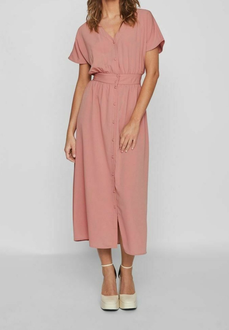 Femme portant une robe midi rose poudré boutonnée avec manches courtes et taille cintrée, associée à des talons compensés beige.