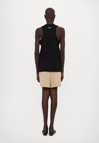 Melnas bezpiešu topiņš ar ikdienišķu piegriezumu, bēšas šortus un melnas plakanas kurpes, demonstrējot minimālistisku dizainu ar diskrētu zīmola identitāti aizmugurē.