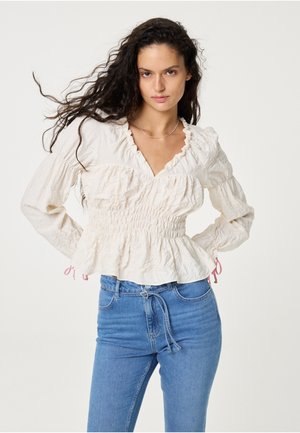 Woman with long dark curly hair wearing a textured cream blouse with ruffles and high-waisted blue jeans, standing with hands on hips.