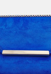 Bolso de mano azul de ante con un detalle metálico plateado pulido y una cadena trenzada en la parte superior. Textura suave con un color vívido.
