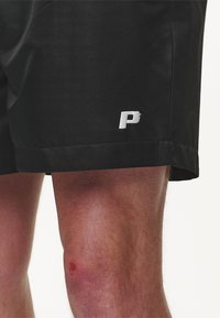 Pantaloni corti neri realizzati in un tessuto leggero con una texture liscia. Presentano un ricamo bianco a forma di "P" sull'orlo, con la pelle del ginocchio visibile.