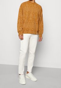 Bluza z okrągłym dekoltem w kolorze musztardowym z tonowanym wzorem, zestawiona z białymi prostymi dżinsami i białymi sneakersami. Prosty design, casualowy krój.