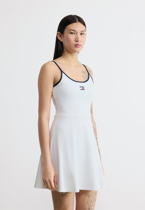 Νέα γυναίκα με μακριά μαύρα μαλλιά που φοράει λευκό αμάνικο φόρεμα με ναυτικό περίγραμμα και μικρό κόκκινο, λευκό και μπλε λογότυπο στο στήθος.