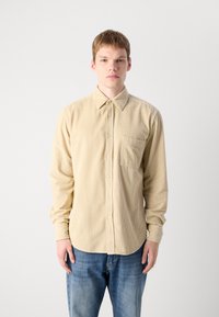 Cămașă bej din corduroy cu guler button-down, buzunar în față și croială dreaptă. Mânecilor lungi au manșete cu nasturi.