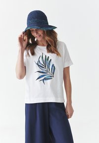 T-shirt en coton blanc avec un motif de feuilles vertes et bleues sur le devant, associé à un pantalon large bleu marine et un chapeau tissé bleu.
