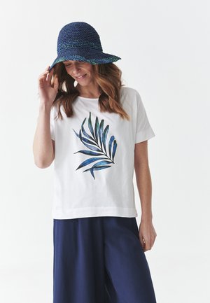 T-shirt en coton blanc avec un motif de feuilles vertes et bleues sur le devant, associé à un pantalon large bleu marine et un chapeau tissé bleu.