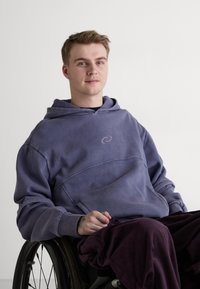 Lilla hettegenser med en glatt tekstur, med en subtil virvel-logo. Mørkelilla corduroy bukser, sittende i rullestol.