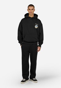 Czarna bluza z kapturem, z kieszenią kangurką, z okrągłym wyszywanym logo. W połączeniu z fakturowanymi czarnymi spodniami i czarnymi sneakersami.