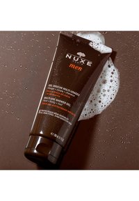 NUXE:s multifunktionella duschgel för män i en mörkbrun tub, med orange detaljer och en strukturerad etikett, omgiven av tvålskum och vattendroppar.