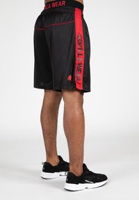 Zwarte shorts met rode zijaccenten, voorzien van "GORILLA WEAR" in verticale print. Satijnen afwerking, elastische tailleband, en een ritssluiting pocket aan de zijkant.