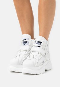 Adidași înalți albi din piele netedă, cu guler căptușit, accente cu logo, tălpi groase din cauciuc și curele Velcro.