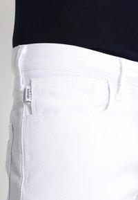 White denim trousers featuring a straight leg, visible stitching, and a label on the pocket. The waistband is smooth and elasticized.
