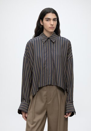 Femme aux longs cheveux foncés portant une chemise ample à rayures foncées boutonnée et un pantalon marron large à taille haute, debout devant un fond uni.