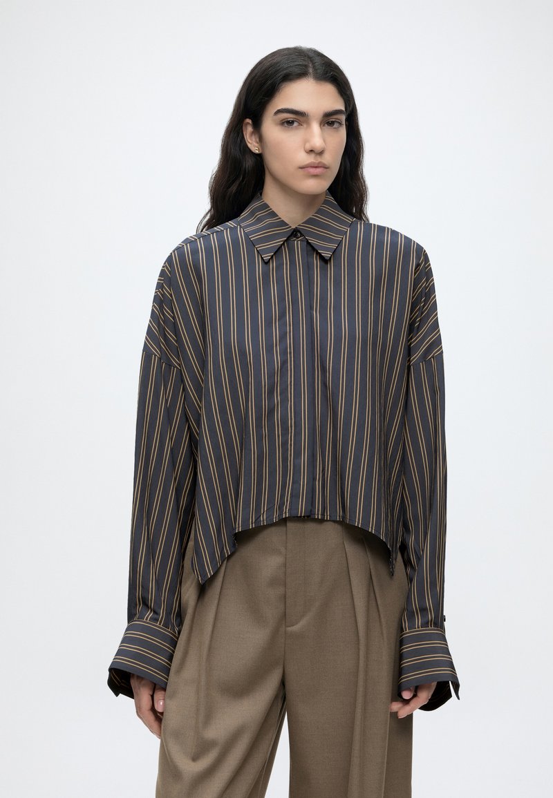 Femme aux longs cheveux foncés portant une chemise ample à rayures foncées boutonnée et un pantalon marron large à taille haute, debout devant un fond uni.