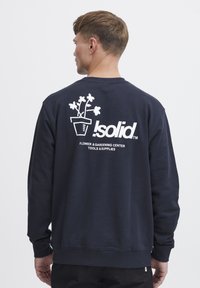 Felpa navy con collo a giro, caratterizzata da una grafica bianca di un vaso di fiori e il testo "Isolid Flower & Gardening Center" sul retro.