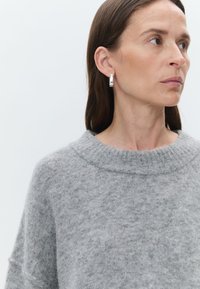 Suéter de lã cinza com um decote largo e punhos canelados. Modelo a usar um brinco de argola prateada, apresentando uma textura lisa e um design geométrico.