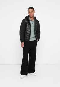Sort pufferjakke med en blank overflade og hætte, båret over en lys grøn polo t-shirt, parret med sorte vide bukser og hvide sneakers.
