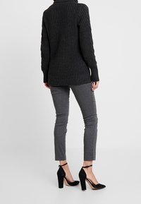 Femme portant un pull en mailles épaisses noir, un pantalon slim gris et des chaussures à talons hauts noires avec bride cheville, se tenant contre un fond uni.