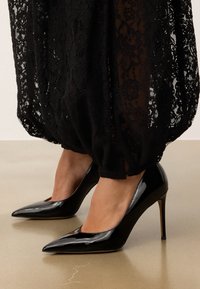 Mustmust mustkonnad kõrged kontsad terava ninaga ja peene stilettokontsaga, mis peegeldavad valgust, koos musta pitsist rõivaga.