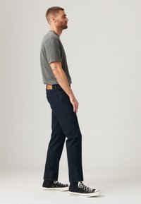 Gray short-sleeve t-shirt paired with dark blue straight-leg jeans and black high-top sneakers. Distinctive tan leather patch on jeans.
