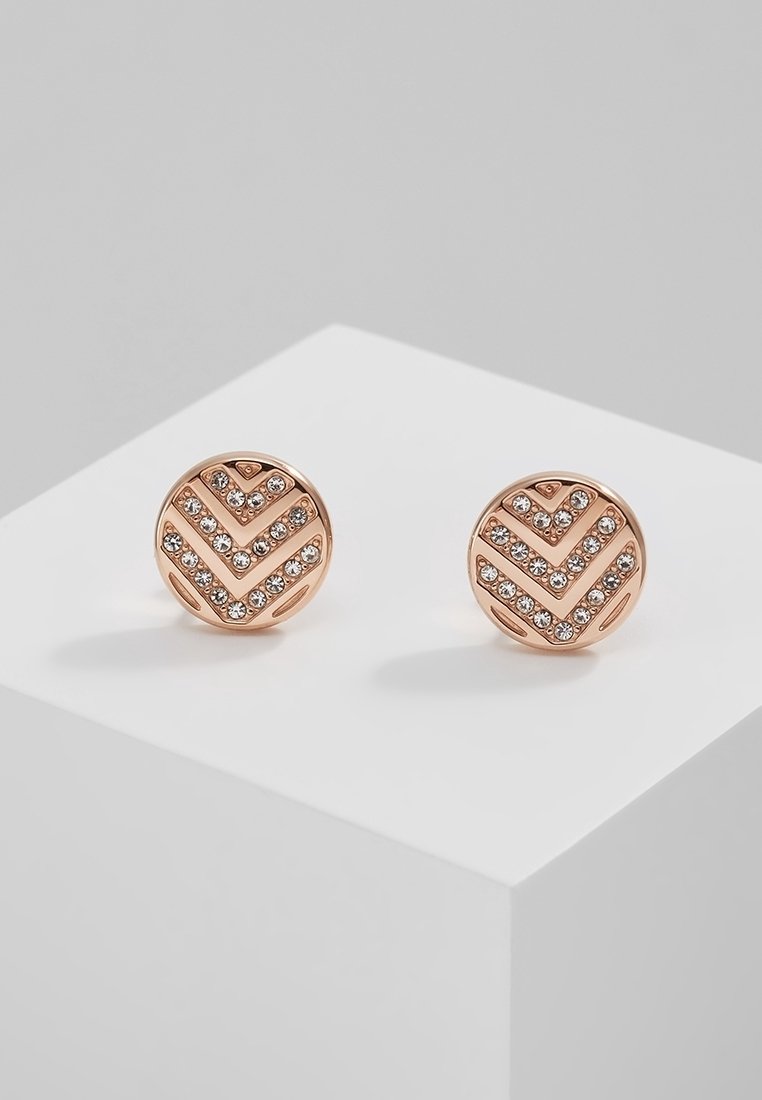 Boucles d'oreilles clous rondes en or rose avec motifs chevrons ornés de petits cristaux clairs sur une surface d'exposition blanche.